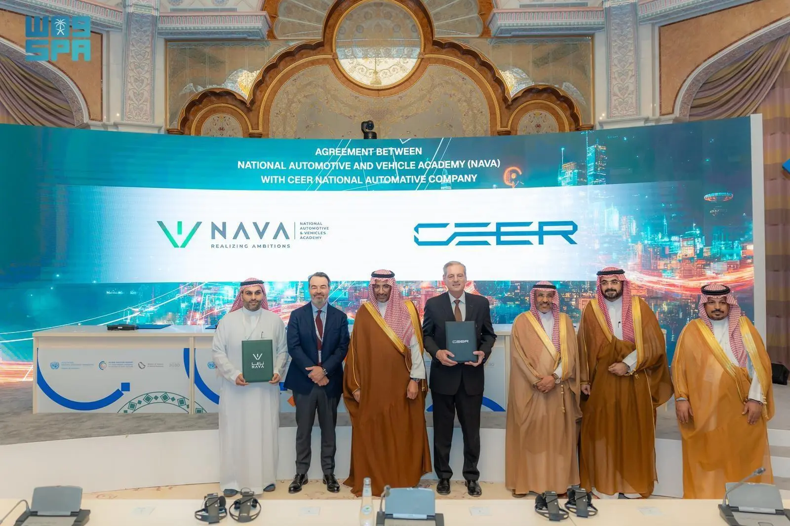 Group of officials from NAVA and CEER National Automotive Company pose on stage after signing a strategic agreement, holding branded folders in front of a large digital screen displaying both logos, Riyadh, Saudi Arabia.
