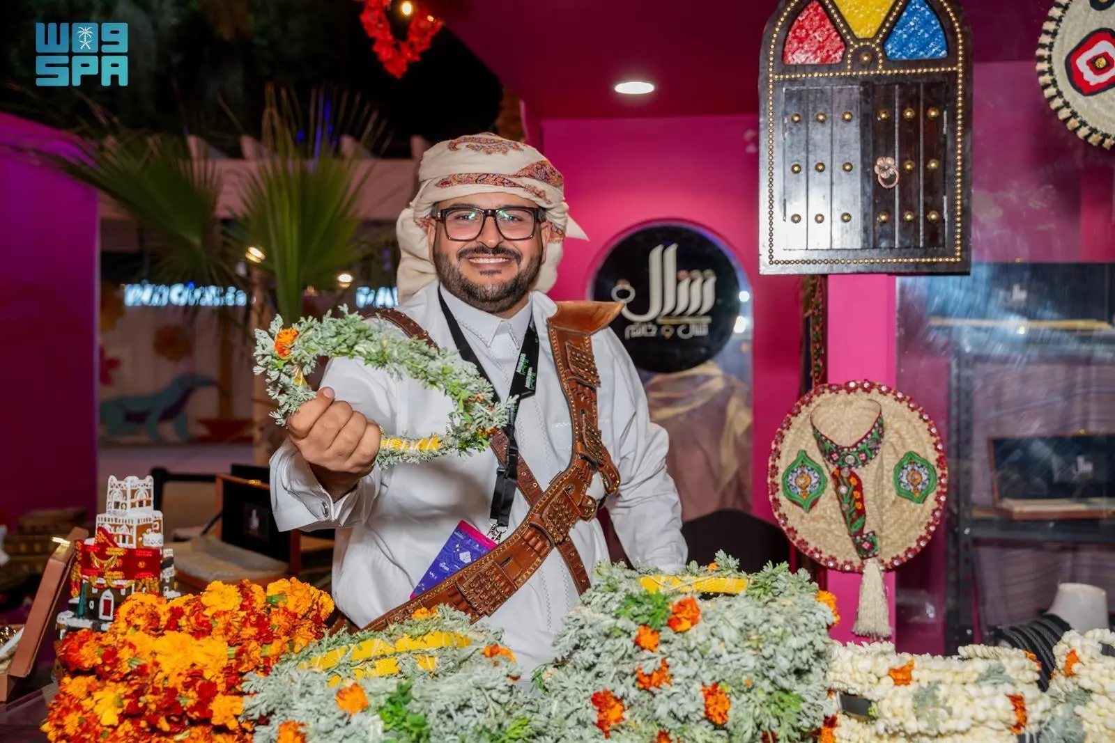 A smiling man wearing traditional Yemeni attire and a janbiya dagger holds up a floral wreath made of marjoram and flowers during Yemeni Cultural Days at the Global Harmony initiative in Riyadh.