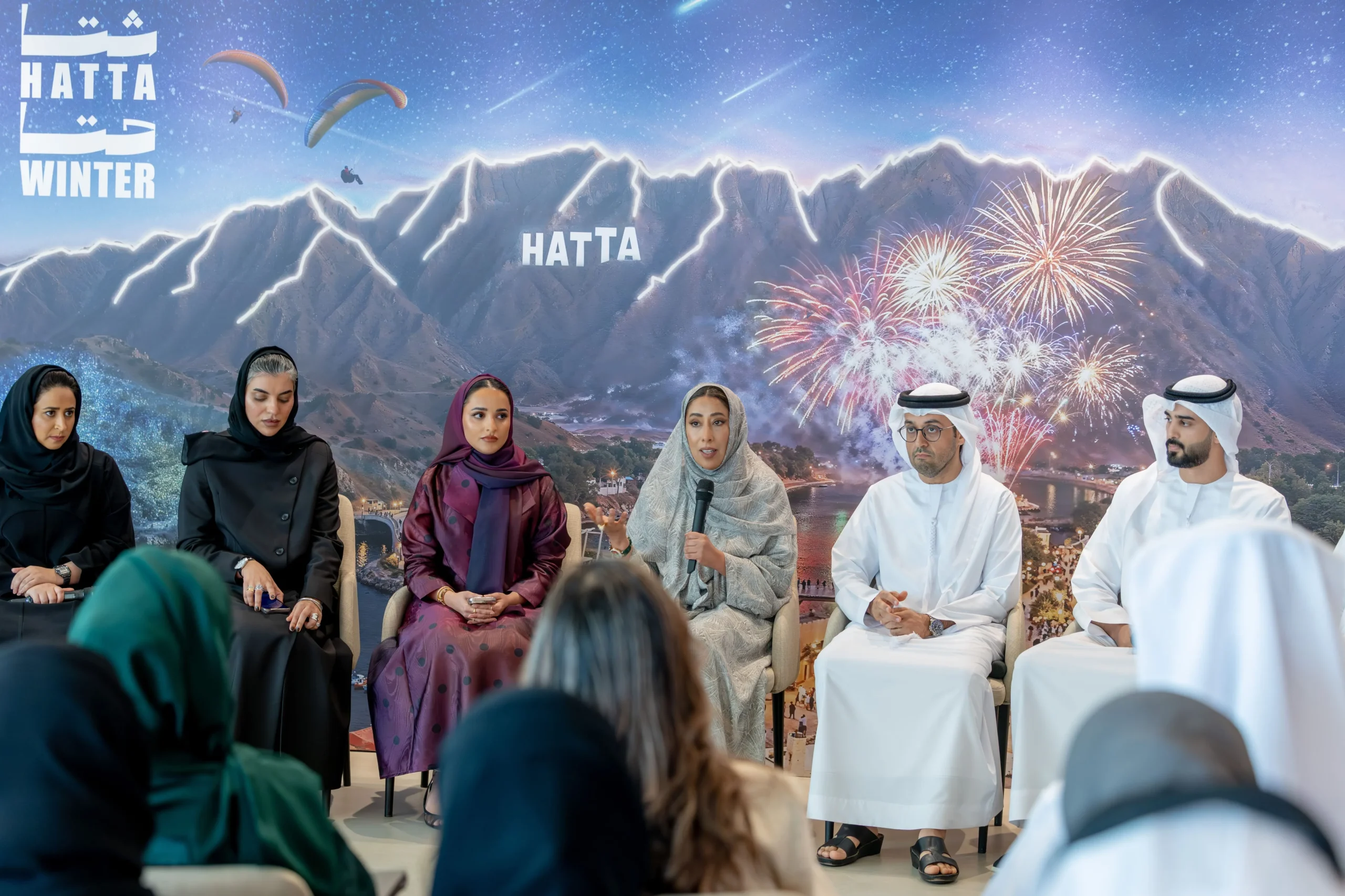Panelists including Mona Al Marri speaking at the press conference announcing the second edition of HattaWinter at the Government of Dubai Media Office, with Hatta mountains and fireworks backdrop.