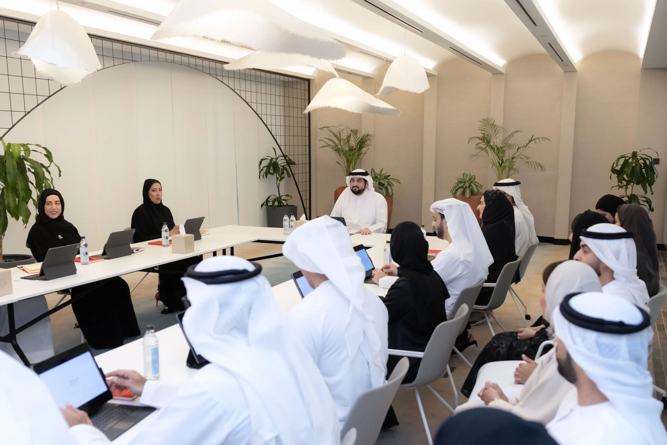 A diverse group of professionals in traditional Emirati attire, including men in white dishdashas and ghutras and women in black abayas and hijabs, seated around a long white conference table in a modern meeting room. Several participants use laptops and tablets, with water bottles and documents on the table. H.H. Sheikh Ahmed bin Mohammed bin Rashid Al Maktoum, wearing a white dishdasha and ghutra, sits centrally at the head, engaged in discussion. The room features contemporary decor with white hanging lamps, potted green plants, beige walls, and a grid-patterned archway.