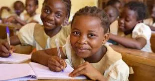 Group of smiling African schoolgirls in beige school uniforms sitting at wooden desks in a classroom, writing in notebooks with pens, focused on learning.