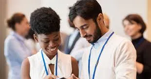 A diverse group of professionals in a conference setting, including a Black woman with short curly hair smiling while looking at a tablet, a Middle Eastern man with a beard leaning in attentively, and several others wearing blue lanyards and business attire, gathered in a modern indoor space with white walls.