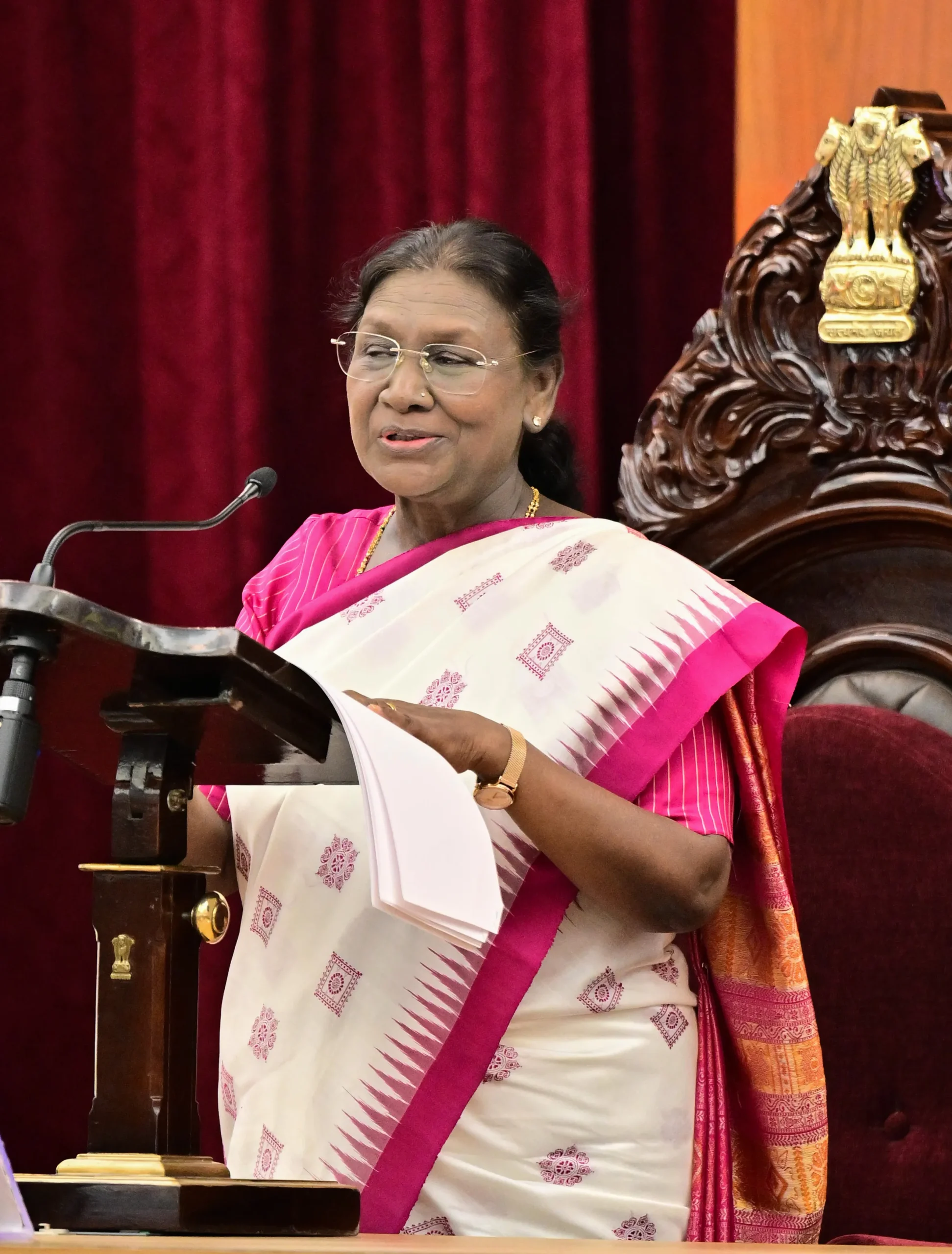 President Droupadi Murmu addressing the Odisha Legislative Assembly at Bhubaneswar on November 27, 2025, standing at the podium in a white and pink saree with the Ashoka Pillar emblem visible behind her.