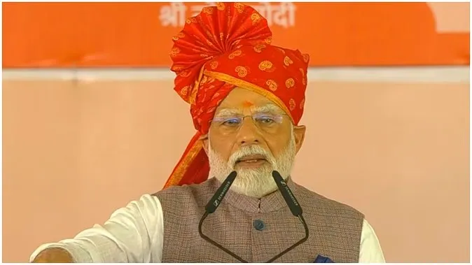 Prime Minister Narendra Modi wearing a red traditional turban with golden patterns, a grey Nehru jacket and white kurta, speaking at a public event with a saffron backdrop displaying Hindi text "श्री नरेंद्र मोदी".