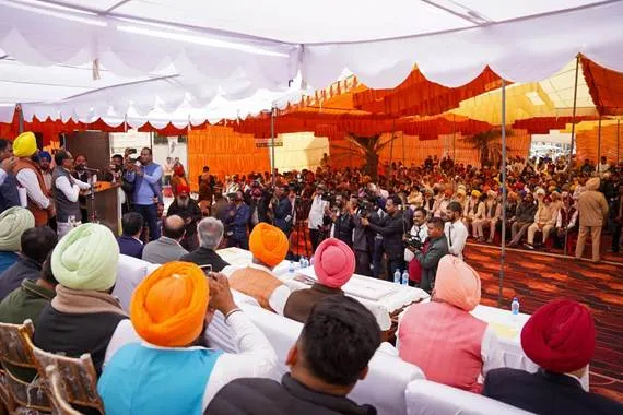 A large outdoor gathering under a white and orange tent in Ransih Kalan village, Moga, Punjab, where Union Agriculture Minister Shivraj Singh Chouhan, wearing a saffron turban and vest, sits at a central table with local officials and farmers in traditional Punjabi attire including colorful turbans and kurtas, addressing the crowd seated on red carpeted floor amid banners and water bottles.