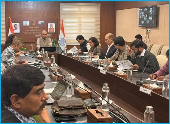 A formal conference room with a long wooden table holding laptops, documents, water bottles, microphones, and notepads. Officials in suits, kurtas, and sarees, including men and women of various ages, sit attentively around the table, some taking notes or speaking. Flags of India and the Department of Drinking Water and Sanitation are displayed, along with portraits of Prime Minister Narendra Modi. A television screen shows a virtual meeting interface, and beige curtains frame the wooden-paneled walls.