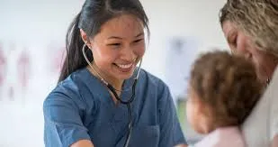 A smiling young Asian female nurse in blue scrubs uses a stethoscope to examine a young child held by an adult patient in a hospital or clinic setting in New Zealand.