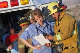 A female paramedic from Hato Hone St John in a light blue uniform consults a clipboard with a Fire and Emergency New Zealand firefighter in full yellow turnout gear and helmet during an emergency response incident in New Zealand.