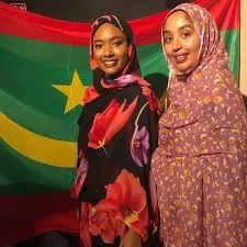 Two Mauritanian women wearing colorful hijabs and traditional clothing smile while standing in front of the national flag of the Islamic Republic of Mauritania during celebrations for the 65th Independence Day.