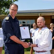 Mark Hamilton OBE, former Deputy Chief Constable of the Police Service of Northern Ireland, shakes hands with a senior Royal Navy officer while receiving a framed certificate during an outdoor appointment or recognition ceremony.